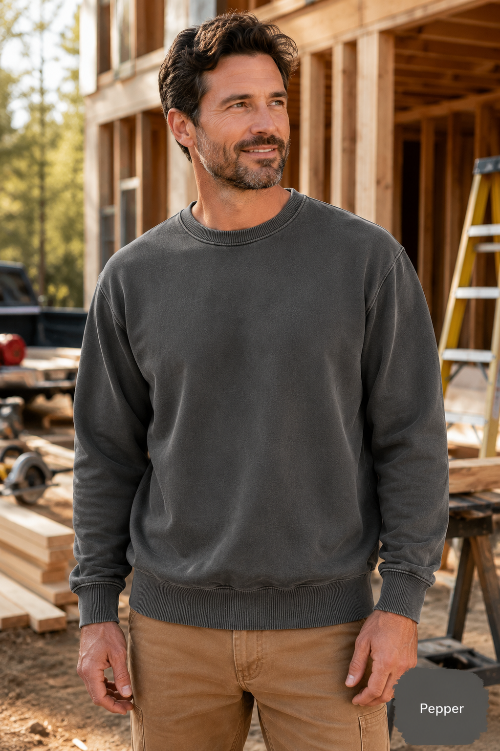 A man wearing a dark charcoal gray crewneck sweatshirt and tan pants, standing in front of a wooden house under construction. A small label in the bottom right corner reads 'Pepper'.