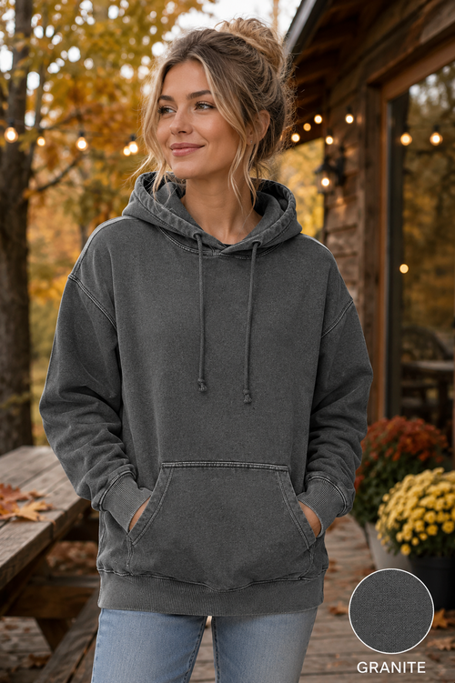 A woman wearing a charcoal gray washed pullover hoodie with a front kangaroo pocket and drawstrings, standing outdoors in front of a rustic wooden building with warm string lights. A circular fabric swatch labeled 'GRANITE' appears in the bottom right corner.