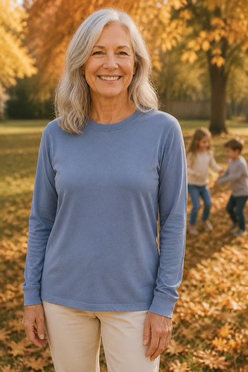 A smiling woman with gray hair stands outdoors in a park wearing a dusty blue long-sleeve crewneck shirt and cream pants, with autumn trees and children blurred in the background.