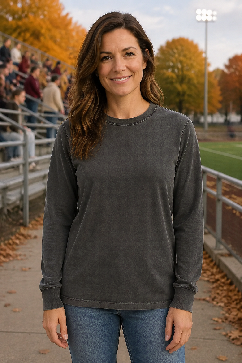 A woman wearing a dark charcoal gray long-sleeve crewneck sweatshirt and blue jeans, standing outdoors on a path at a sports field with autumn trees and stadium seating in the background.