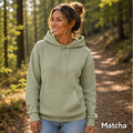 A woman wearing a light sage green ribbed pullover hoodie with a front kangaroo pocket and drawstrings, standing on a forest path. The word 'Matcha' is printed in white text in the bottom right corner.