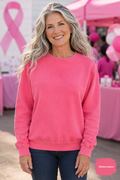 A smiling woman with long wavy gray hair wears a bright pink crewneck sweatshirt and dark blue jeans, standing in front of a pink ribbon tent at what appears to be a breast cancer awareness event.
