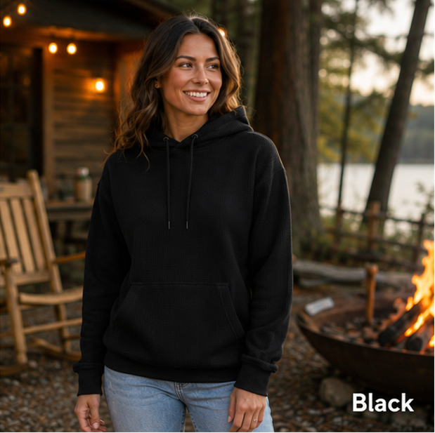 A woman wearing a black pullover hoodie stands outdoors near a campfire and wooden cabin, with the word 'Black' shown in the lower right corner.
