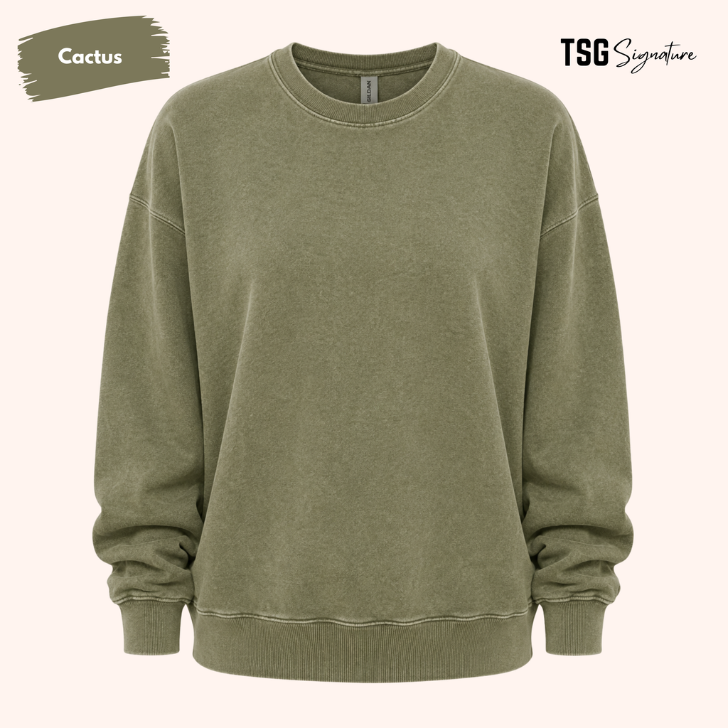 An oversized crewneck sweatshirt in a washed, muted olive green color, labeled 'Cactus' in the top left corner with 'TSG Signature' in the top right, set against a solid pale pink background.