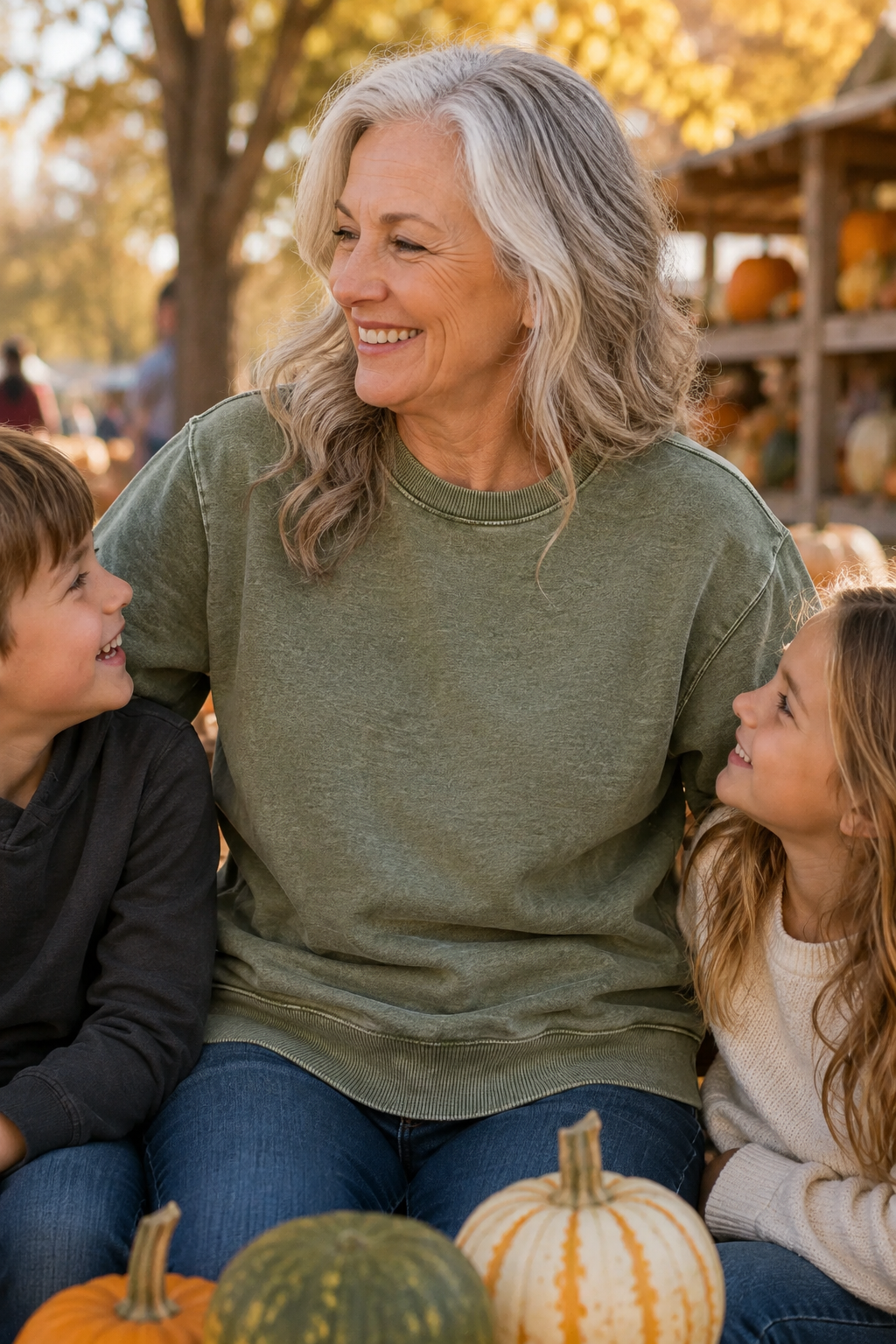 A woman with gray hair wearing an olive green crewneck sweatshirt sits outdoors with two children in a fall-themed setting with pumpkins.