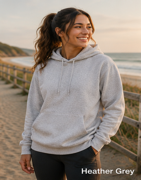 A woman wearing a light heather grey pullover hoodie stands on a sandy beach boardwalk with a wooden fence and ocean in the background. The text 'Heather Grey' is visible in the bottom right corner.