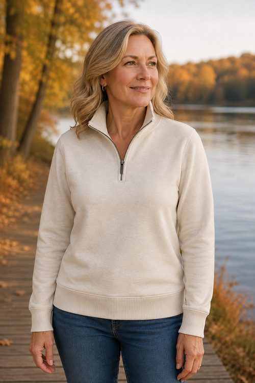 A woman wearing a cream-colored quarter-zip pullover sweater and blue jeans, standing on a wooden boardwalk with a lake and autumn trees in the background.
