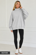 A woman modeling an oversized light heather gray mock neck sweatshirt, paired with black leggings and white sneakers, standing against a white paneled wall. A black label in the bottom-left corner reads 'HEATHER'.