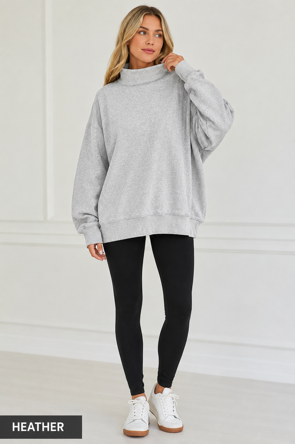 A woman modeling an oversized light heather gray mock neck sweatshirt, paired with black leggings and white sneakers, standing against a white paneled wall. A black label in the bottom-left corner reads 'HEATHER'.