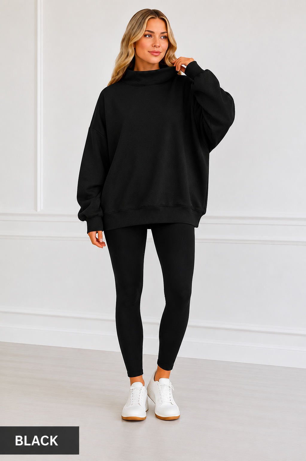 A woman modeling an oversized black sweatshirt with a mock neck, paired with black leggings and white sneakers, standing against a white paneled wall. A black label in the bottom left corner reads 'BLACK'.