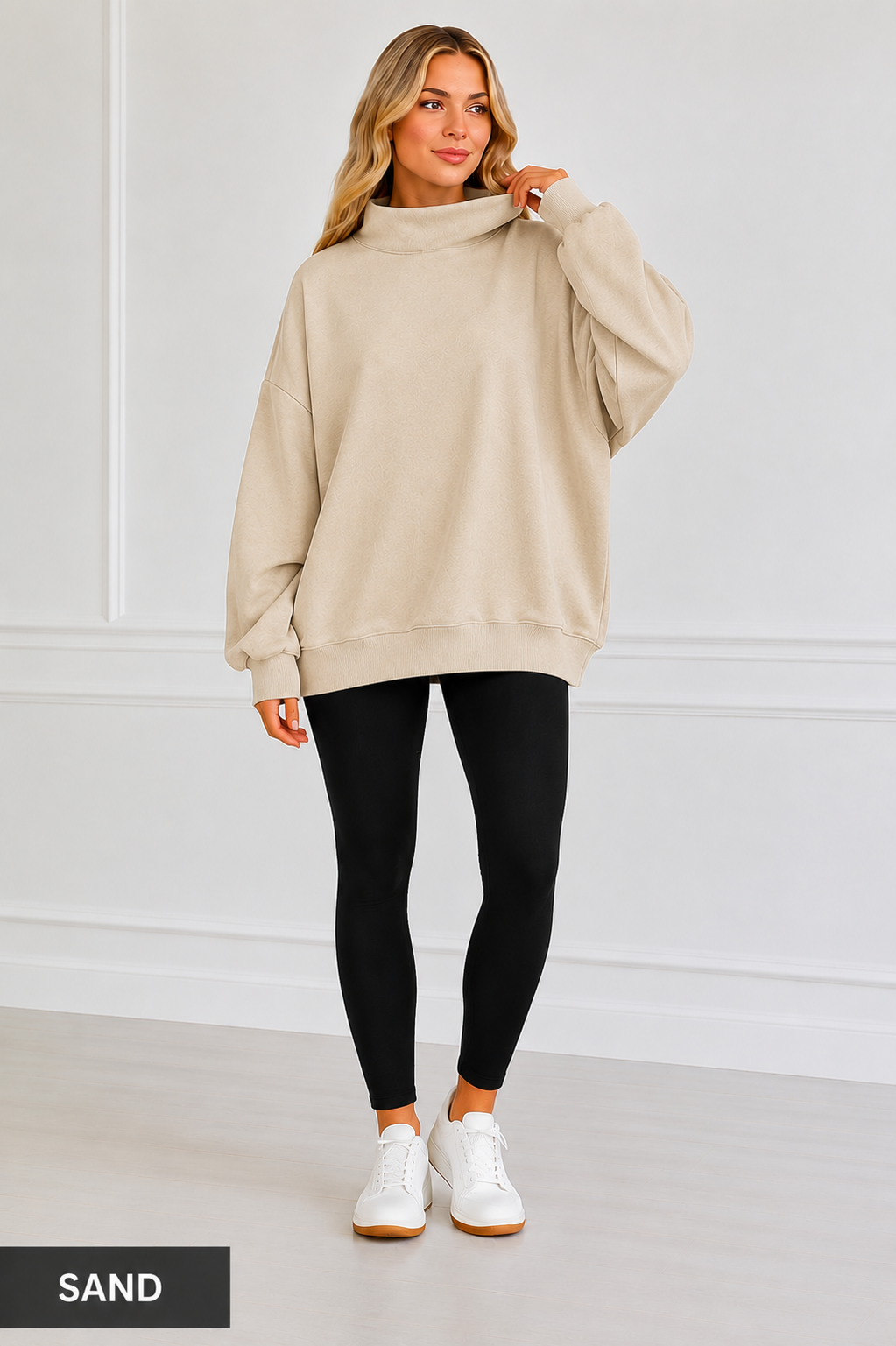 A woman modeling an oversized beige sweatshirt with a mock neck, paired with black leggings and white sneakers, standing against a white paneled wall. A black label in the bottom left corner reads 'SAND'.