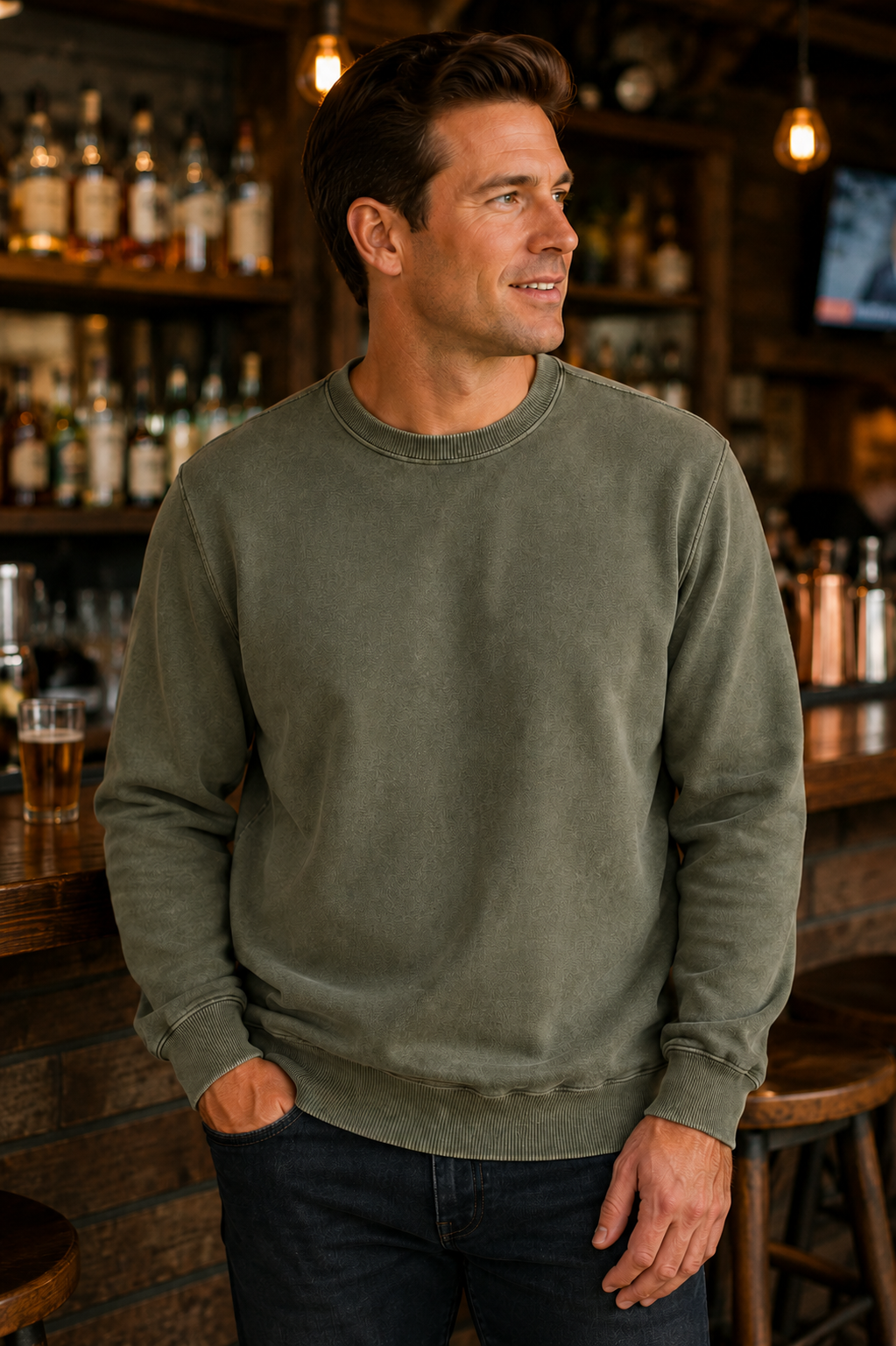 A man wearing a washed olive green crewneck sweatshirt and dark jeans, standing in a warmly lit bar environment.