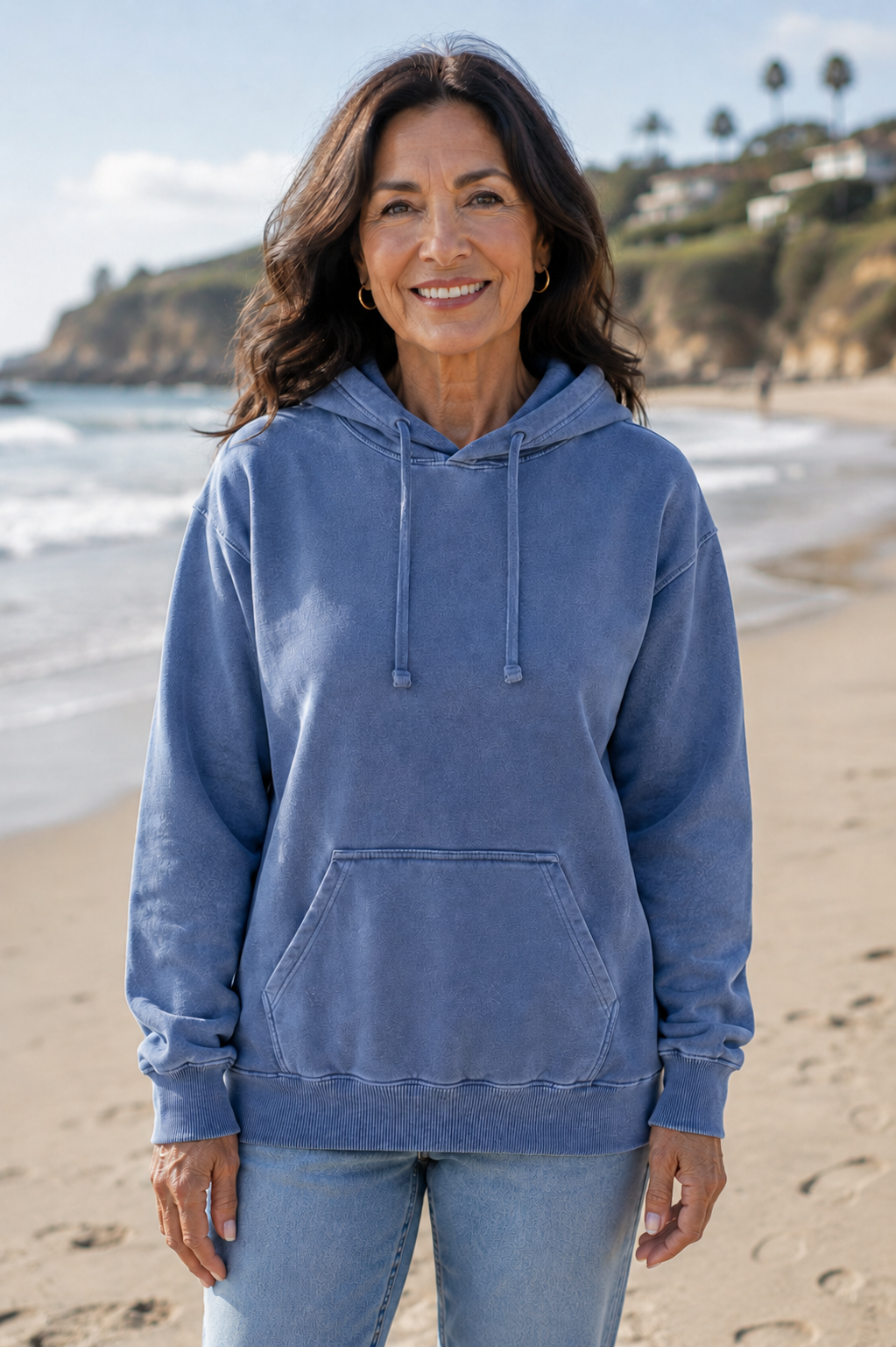 A smiling woman stands on a sandy beach wearing a washed medium-blue pullover hoodie and light-wash jeans, with ocean waves and a rocky coastline in the background.