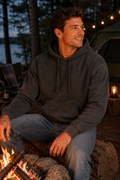 A man wearing a dark charcoal gray washed hoodie and blue jeans sits by a campfire at dusk, smiling and looking to the side. A green tent, string lights, and trees are visible in the background.