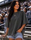 A woman wearing a dark gray oversized short-sleeve t-shirt and denim shorts, standing in a crowded stadium crowd. The word 'SMOKE' is printed on the bottom right corner.