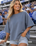 A woman wearing a slate blue oversized short-sleeve top and light denim shorts, standing in a stadium crowd. The brand name 'DUSK' is visible in the bottom right corner.