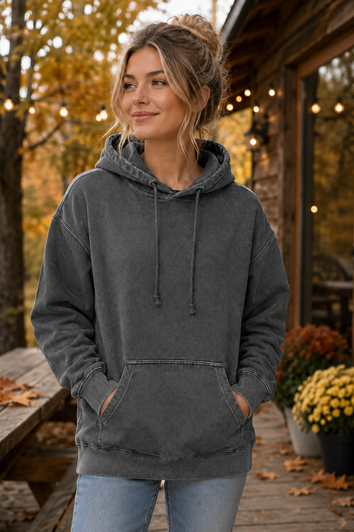 A woman wearing a dark gray washed pullover hoodie with a drawstring hood and kangaroo pocket, standing on a wooden porch in an outdoor autumn setting.