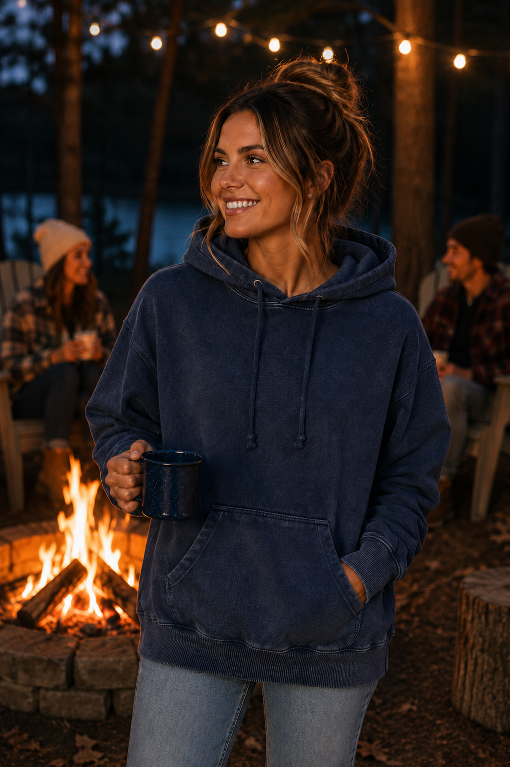 A woman wearing a dark navy blue washed hoodie stands in front of a campfire at night, holding a dark mug. Warm string lights and blurred people sitting in chairs are visible in the background.