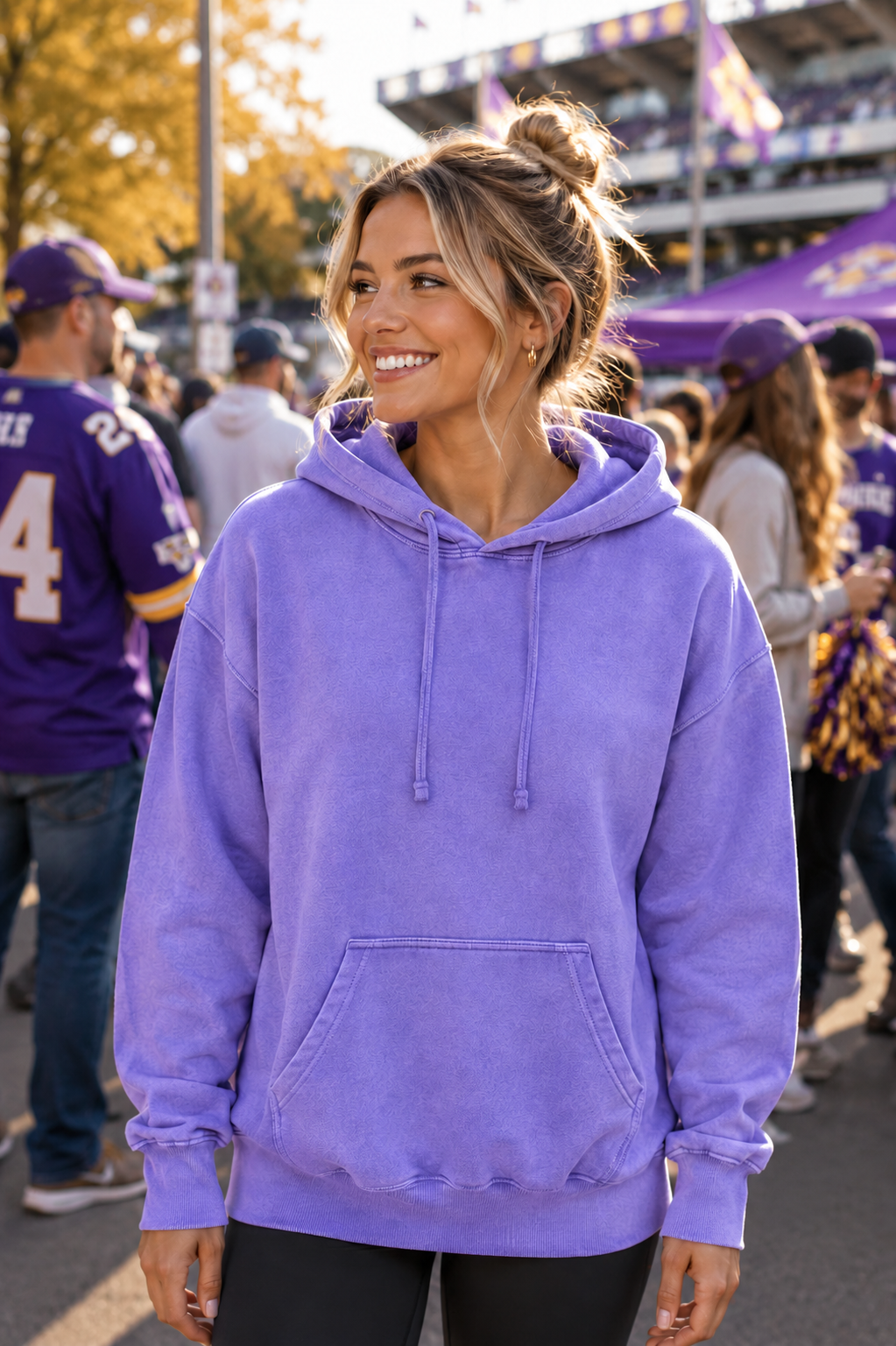 A woman wearing a lavender purple hooded sweatshirt stands at a crowded outdoor event, smiling and looking to the side. The background shows other people, stadium seating, and autumn trees.