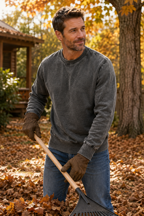 A man in a gray crewneck sweatshirt and blue jeans raking autumn leaves in a backyard with trees and a porch visible in the background.