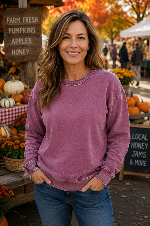 A woman wearing a washed mauve-purple crewneck sweatshirt and blue jeans stands at an outdoor autumn farmers market with pumpkins, flowers, and fall foliage in the background.